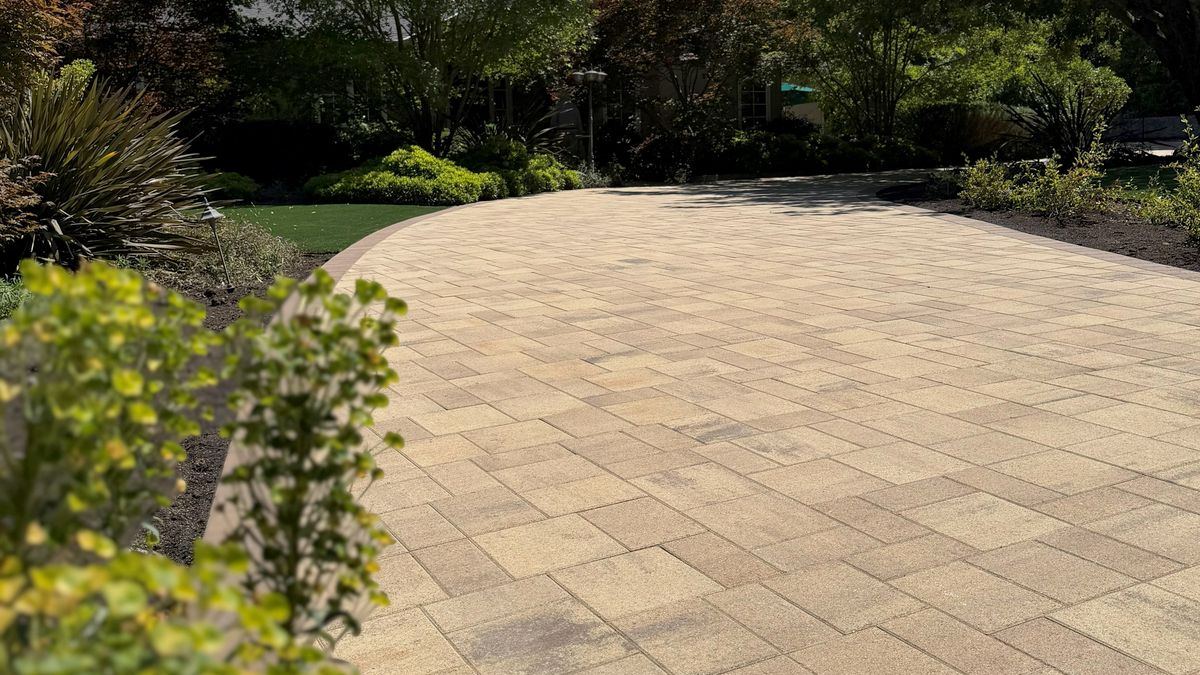 Curved paver driveway in warm sand tones beside planting beds and an artificial turf lawn