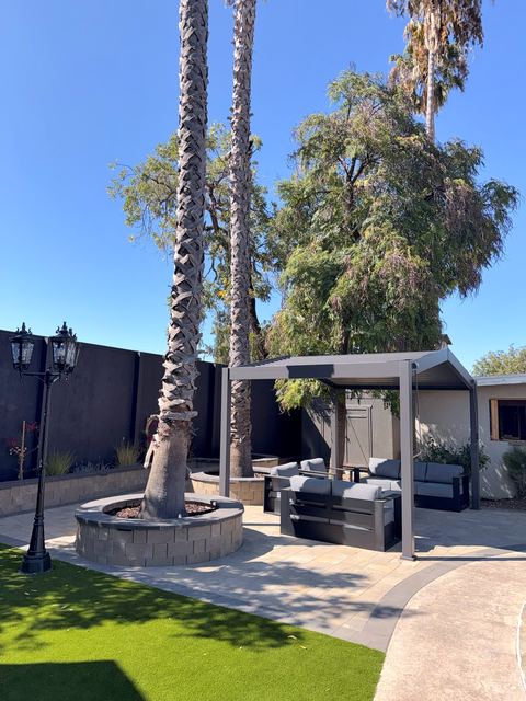 Backyard patio with a palm tree in a round stone seat wall, a lamp post, a pergola and artificial turf