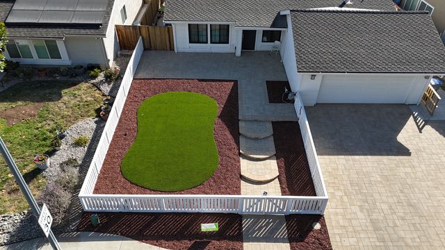 Aerial view of a front yard with a white picket fence, an artificial turf island, red mulch and paver steps