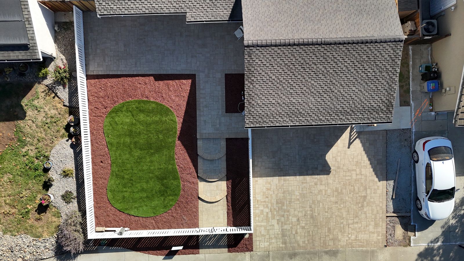 Overhead view of a fenced front yard with an artificial turf island in red mulch, curved paver steps and a paver walkway beside the driveway