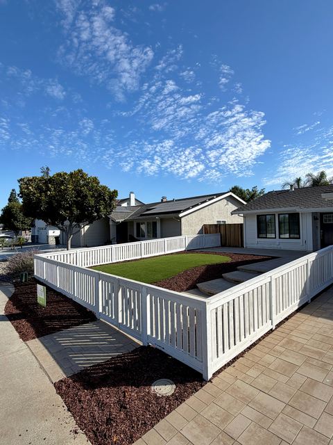 White picket fence around a front yard with artificial turf and curved paver steps, under a blue sky