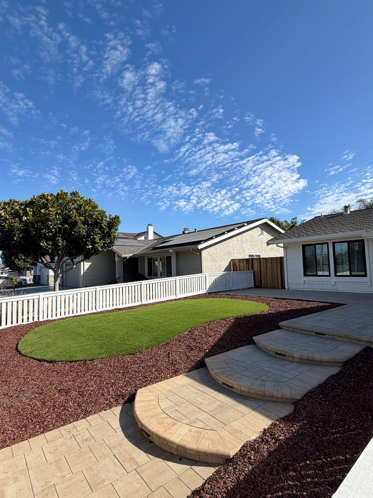 Front yard with an artificial turf island, red mulch and curved paver steps beside the white picket fence